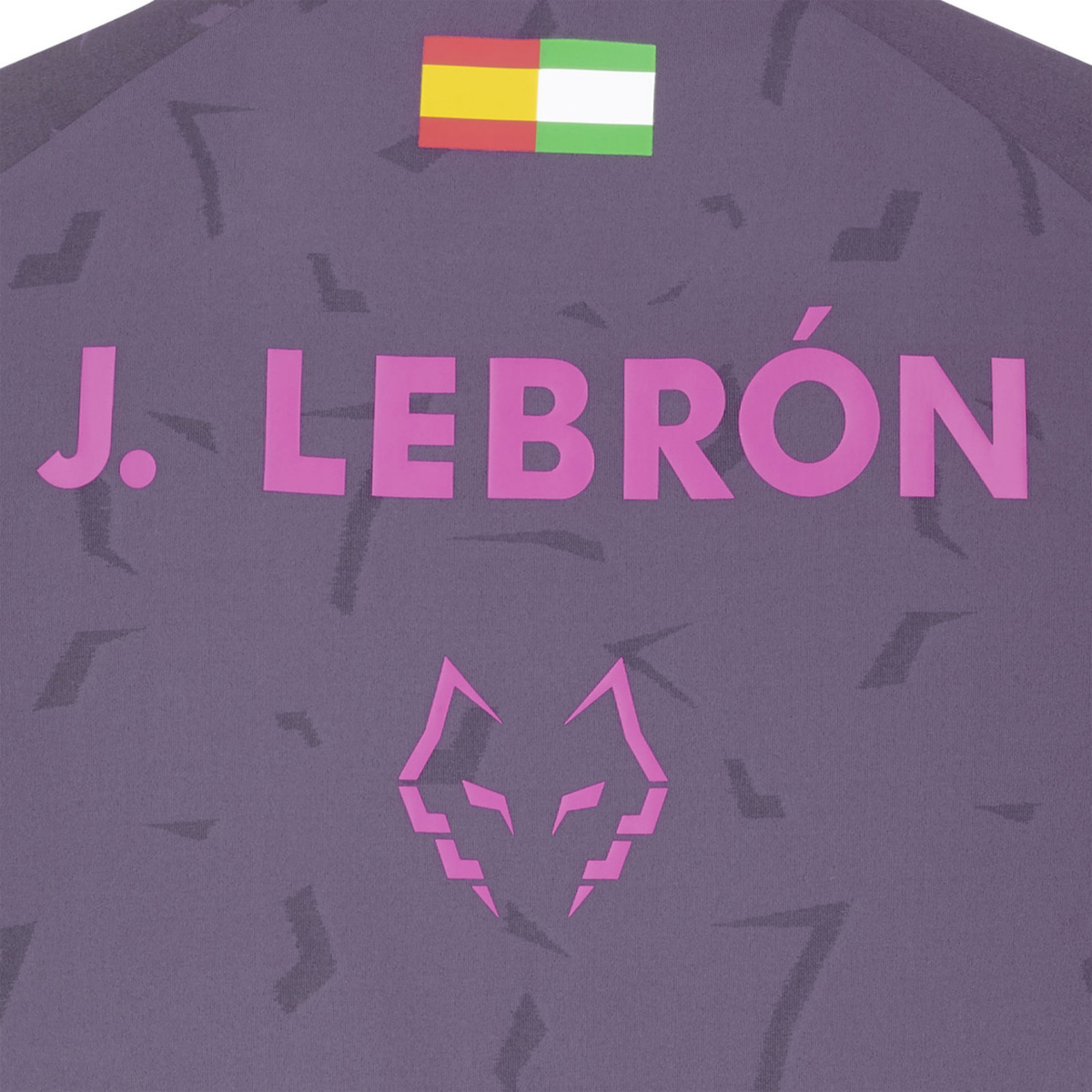 Close-up of the back of the Babolat Juan Lebrón Replica Tee, showing the large pink J. Lebrón lettering, small multicolor flag detail and pink wolf logo on the purple patterned fabric.